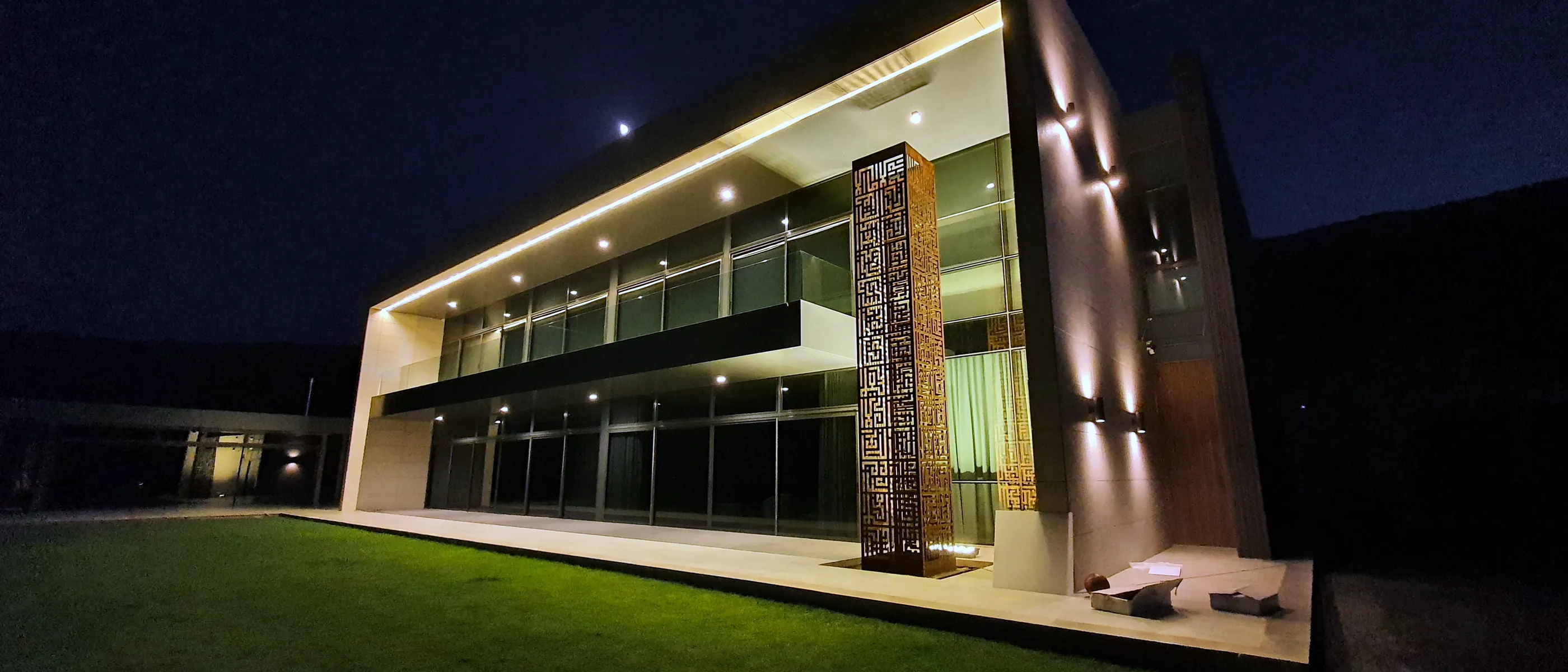 Villa Bhamdoun photographed at night: a long two-storey volume with continuous glazing lit from within, a tall perforated metal screen marking the entrance, and lawn in the foreground.